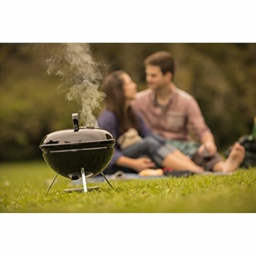 Couple sitting on grass near a smoking barbecue grill.