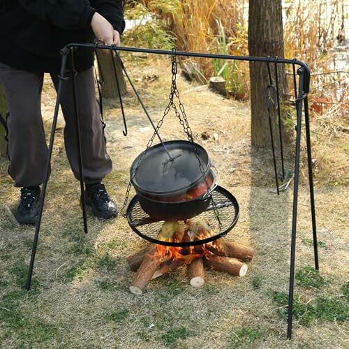 Person cooking over a campfire with a hanging pot and tripod setup.