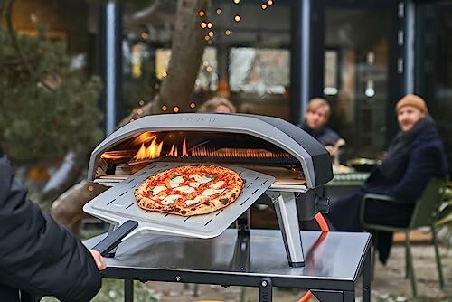 Person using an outdoor pizza oven with a pizza inside, people sitting in the background.